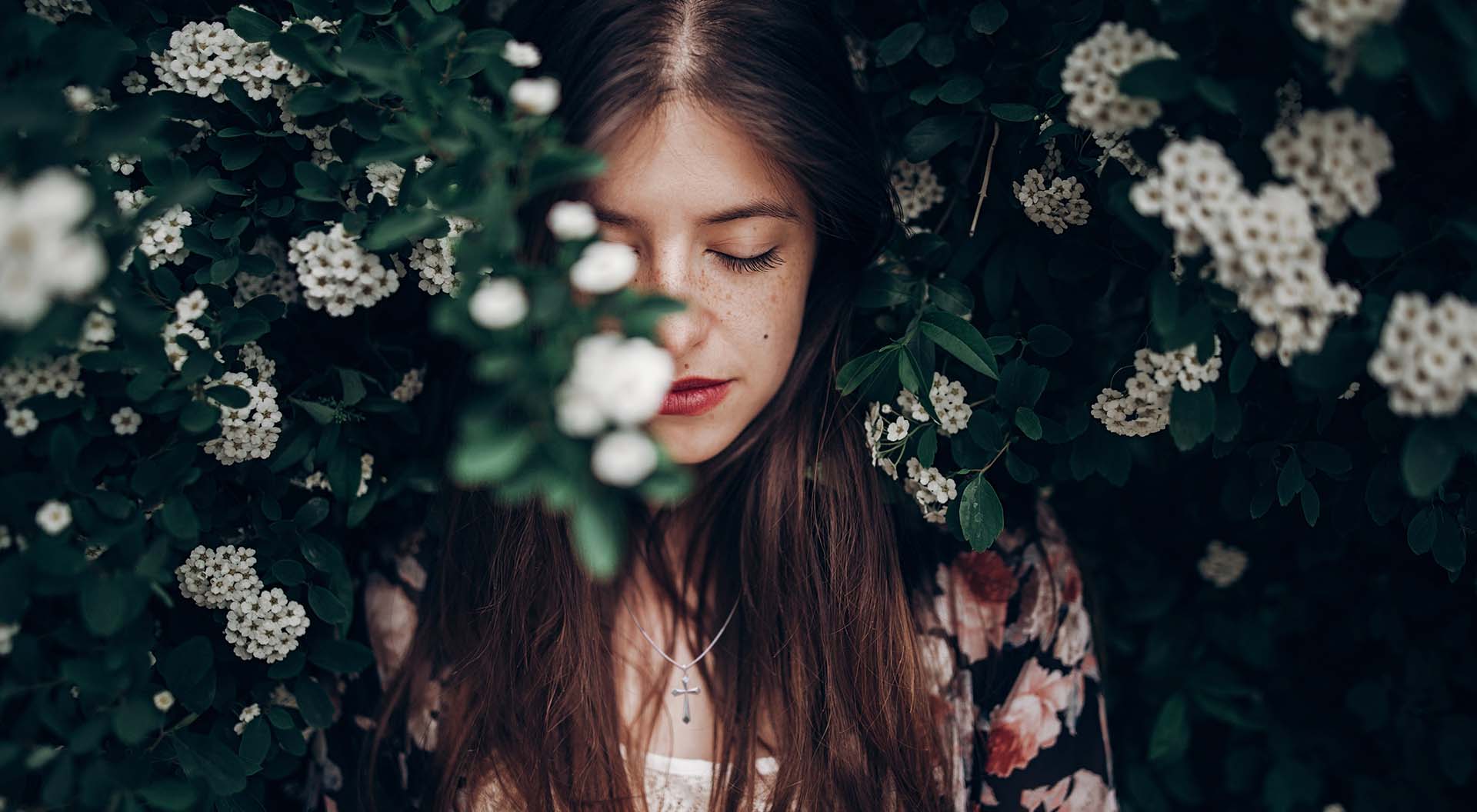 A young woman with her eyes closed, photographed near a flowering bush, with a crucifix around her neck